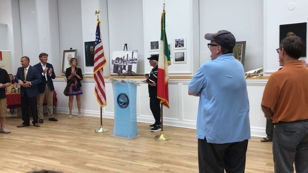 Man in black hat and coat at podium people clapping in background